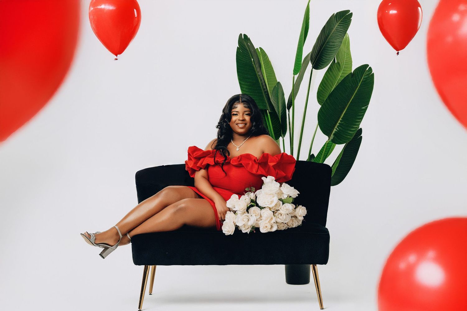 Woman in red ruffled dress reclining on black chair with white roses and red balloons.