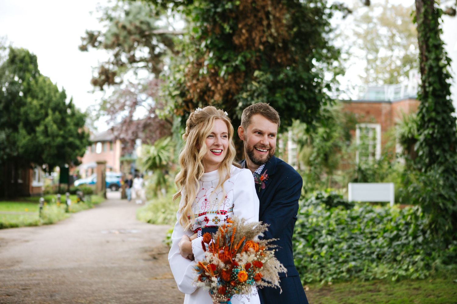 Happy couple laughs while holding autumn-colored wedding bouquet on garden path.