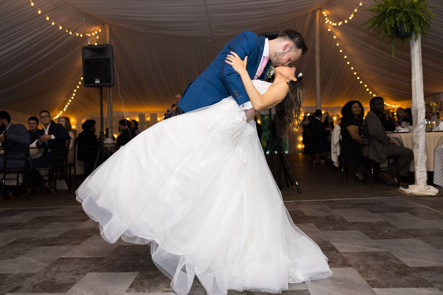 A couple shares their first dance at an evening wedding reception under a white tent with twinkling lights.