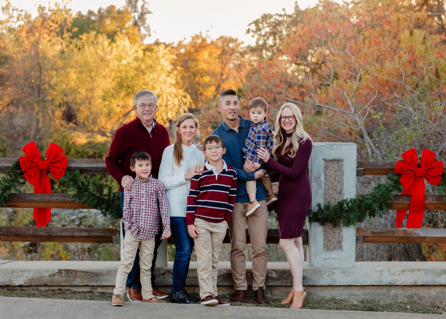 A group stands together for a holiday portrait in front of fall foliage with red bows and evergreen garland decorations.