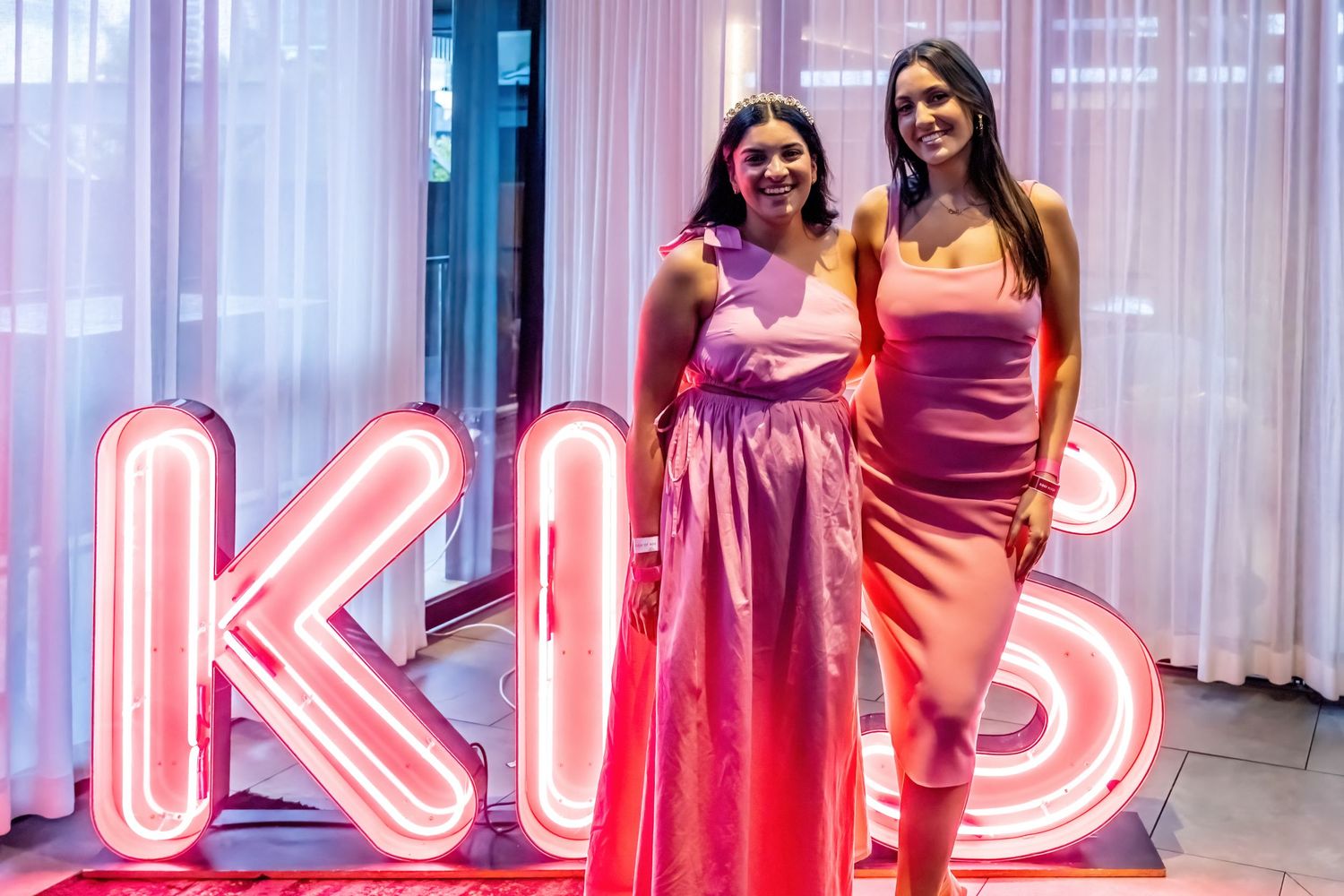 Two women in pink and coral formal dresses pose in front of illuminated KIS neon sign.