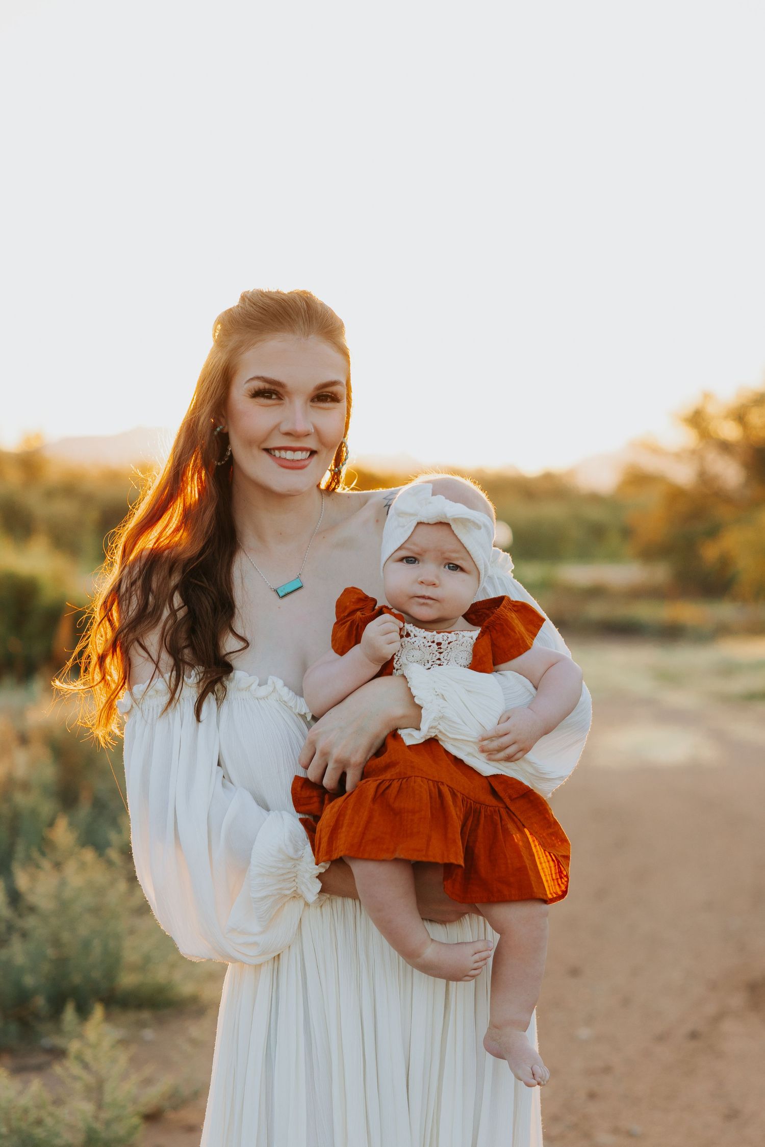 A mother and baby in white and rust-colored outfits pose together in golden sunlight during sunset.