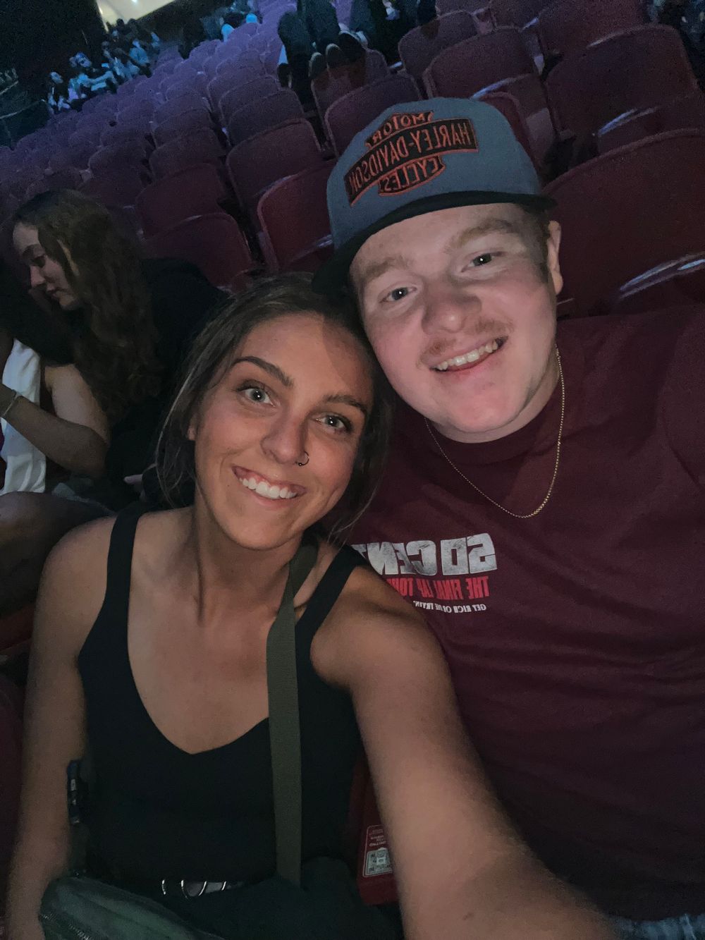 Two people taking a selfie together while sitting in theater seats at an indoor venue.