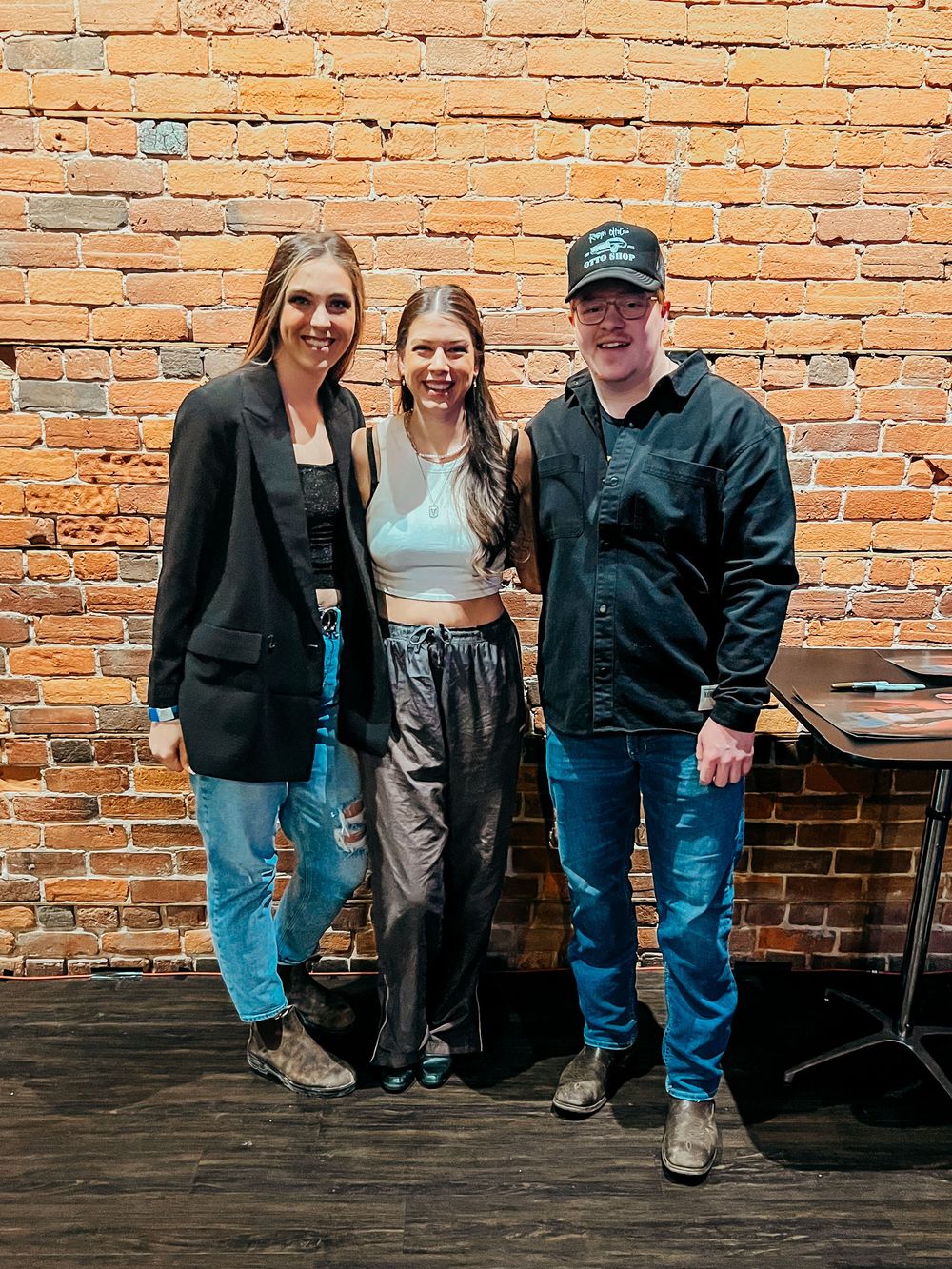 A group stands together posing for a photo against a rustic exposed brick wall in a casual indoor setting.