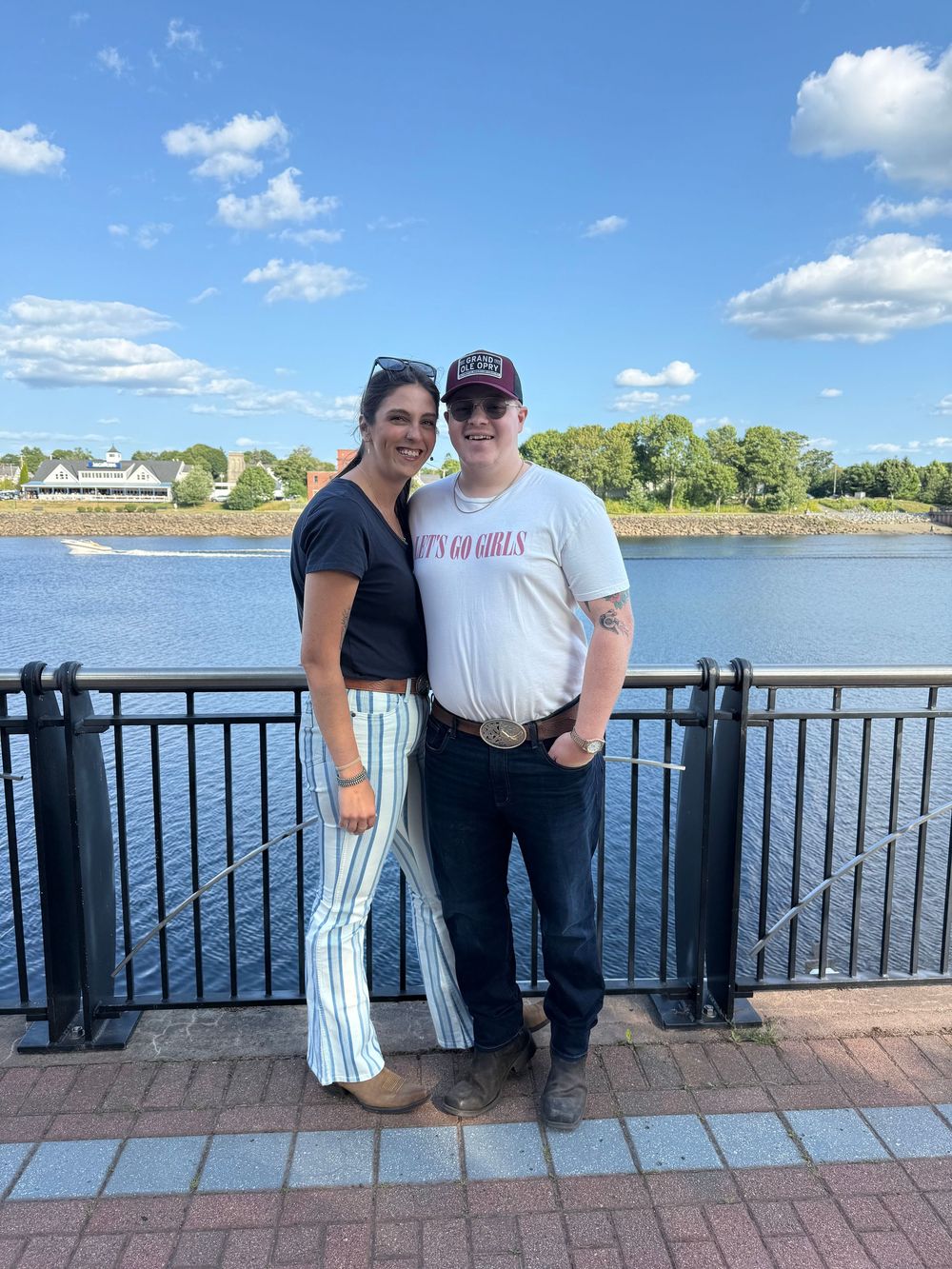 Two people stand together on a brick boardwalk overlooking a scenic waterfront with blue sky and puffy clouds.