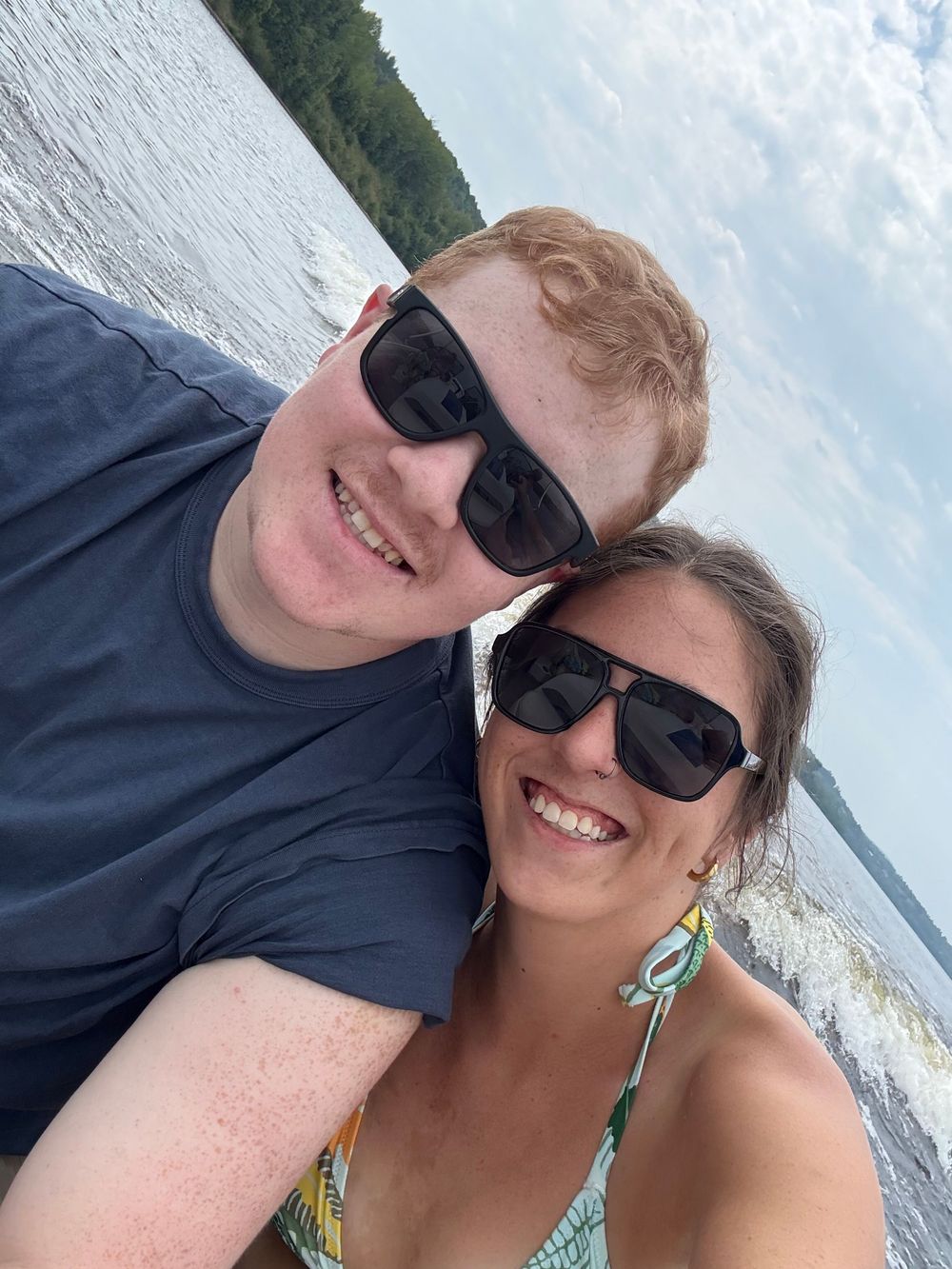 A couple takes selfies together on a boat with cloudy skies and water in the background.