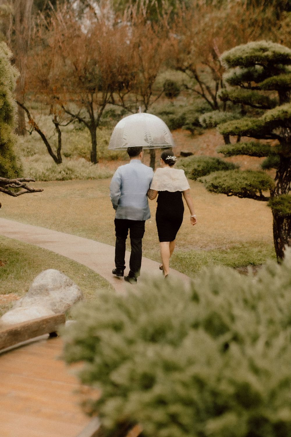 A couple strolls together under a white umbrella on a garden path surrounded by autumn foliage.