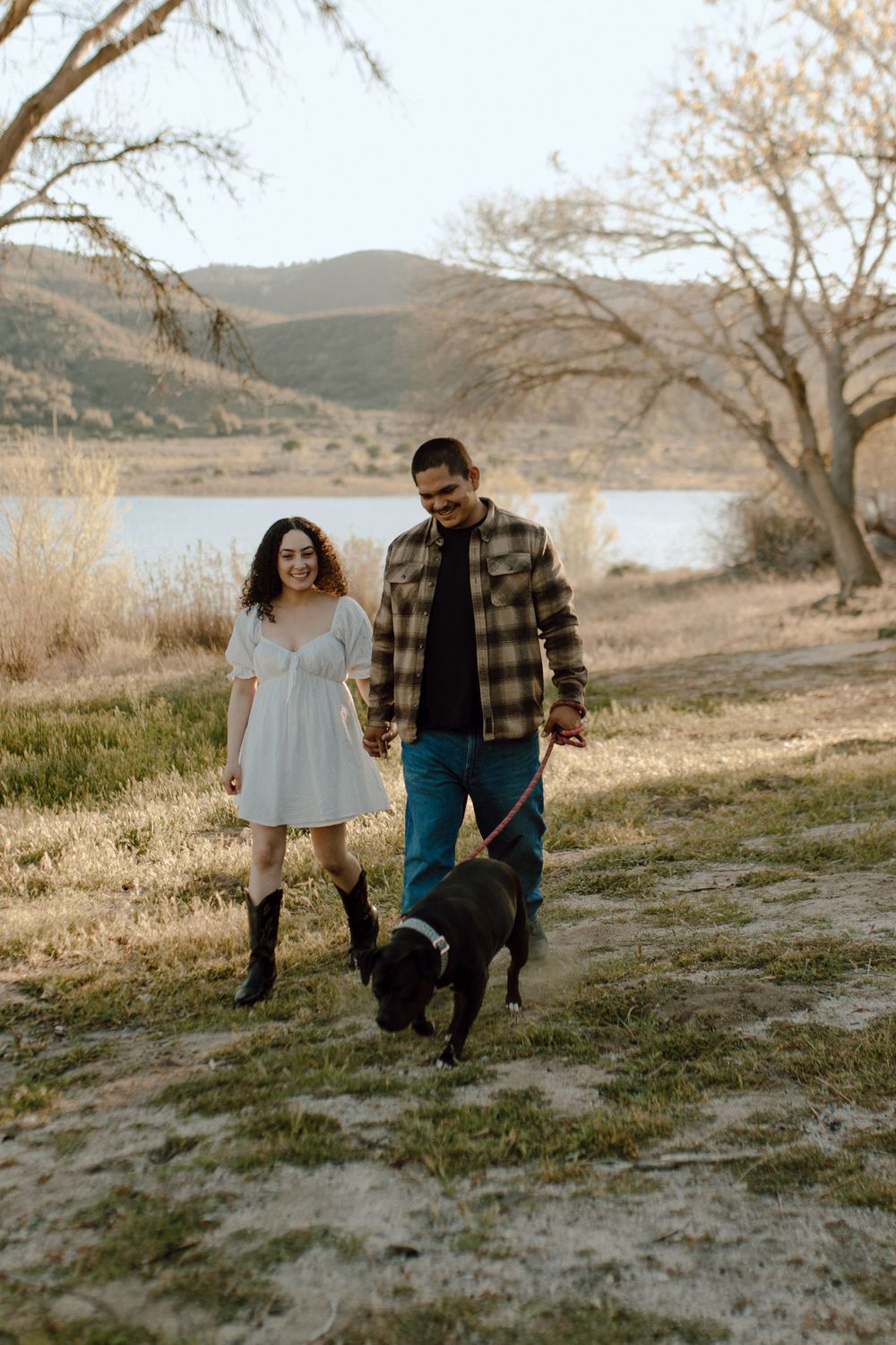 A couple walks their black dog along a lakeside trail during golden hour with mountains in the background.