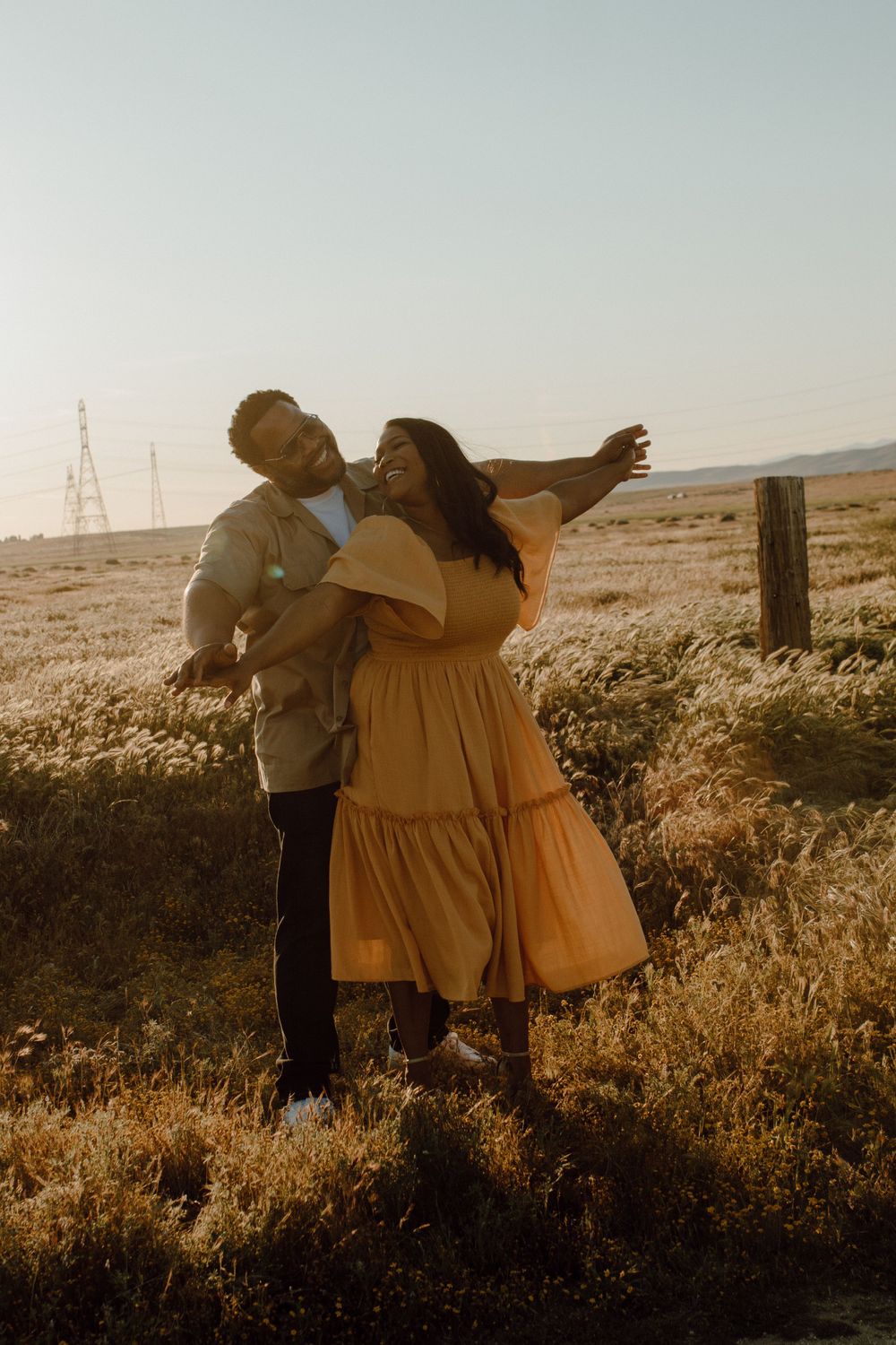 A couple stands together in a golden field at sunset as a yellow dress flows in the breeze.