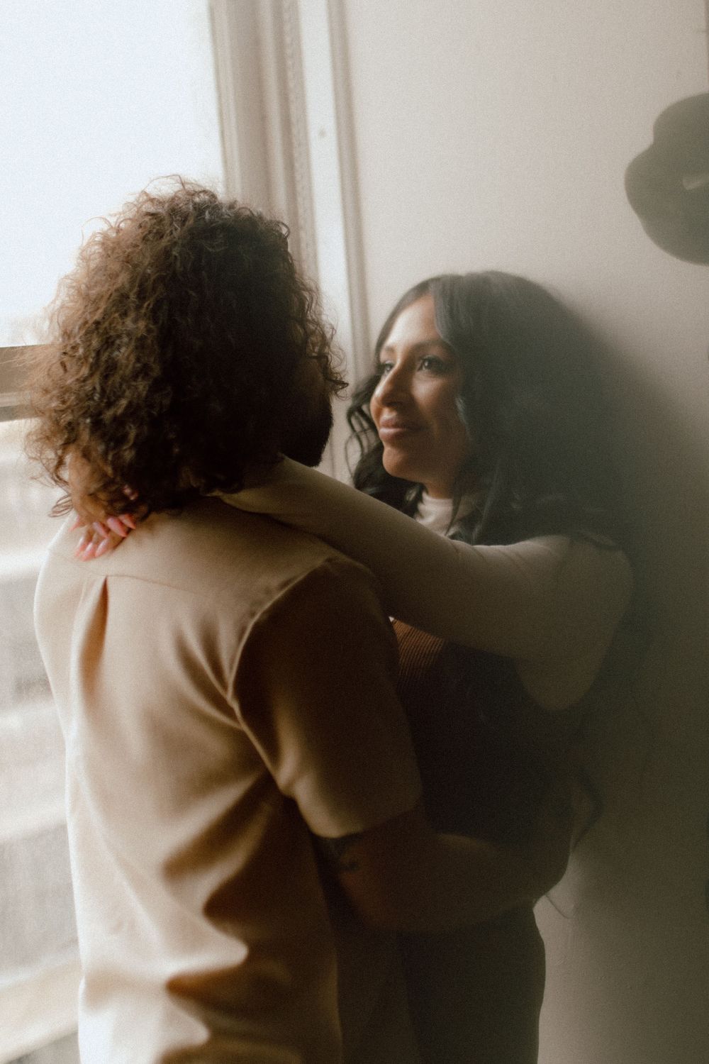 Two people in a tender embrace stand by a window in soft natural lighting, creating an intimate moody atmosphere.