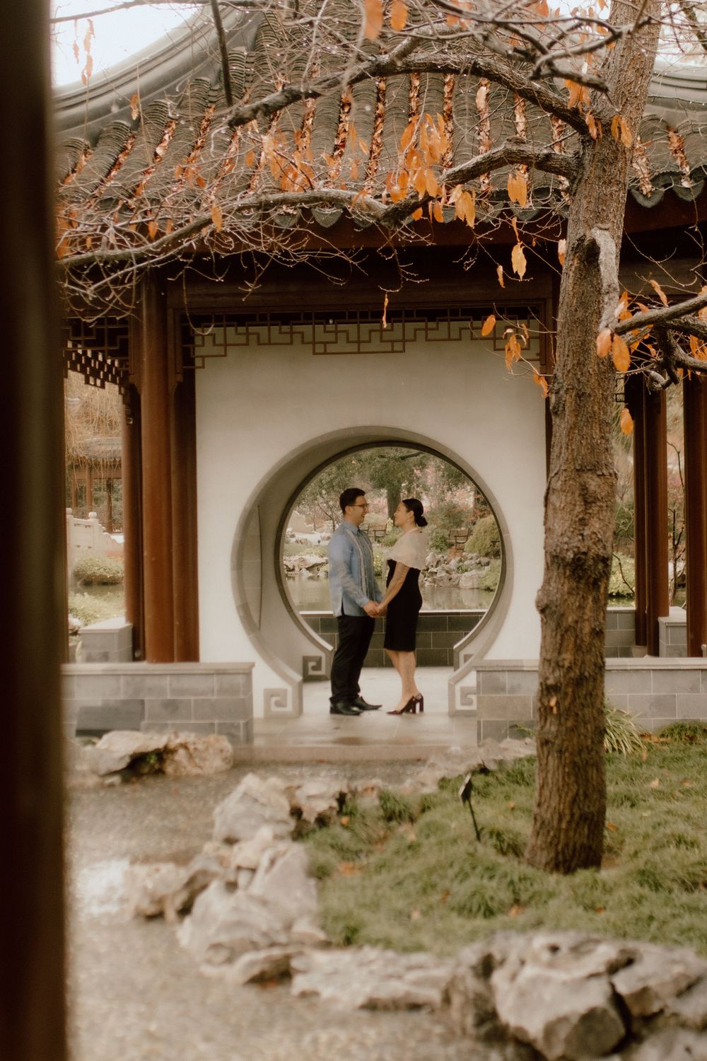 A couple stands in a circular moon gate doorway of a traditional Asian garden structure.