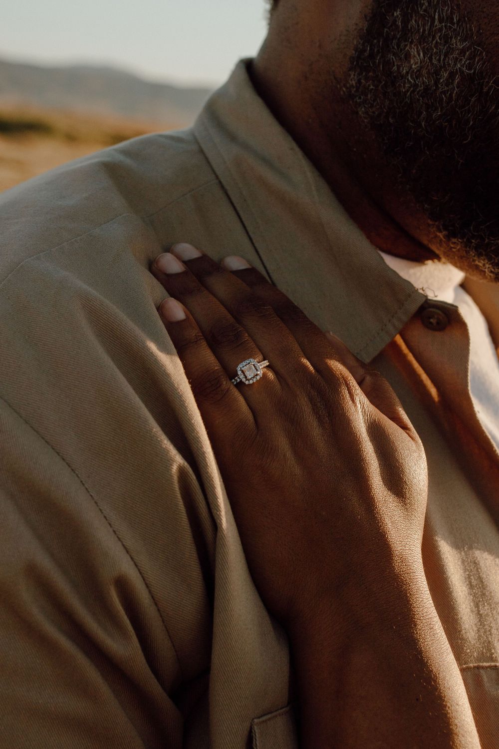 Close-up of a diamond engagement ring worn on a hand against a beige jacket in warm sunlight.