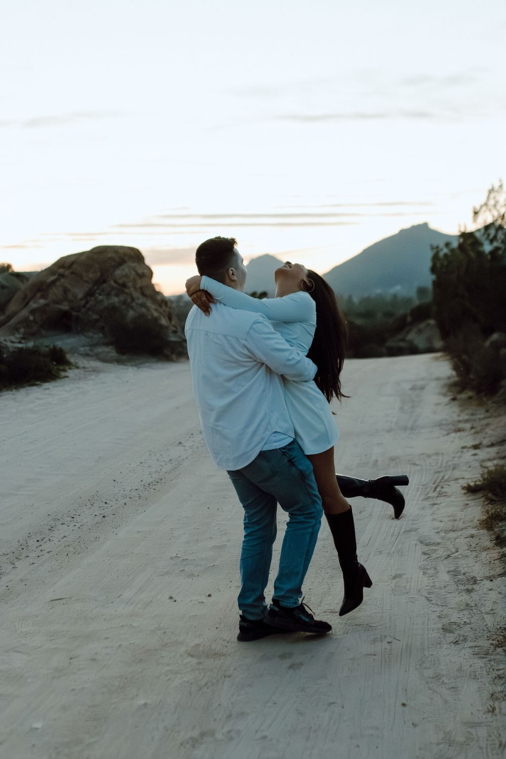A playful couple sharing an intimate moment on a dirt path with mountains in the background.