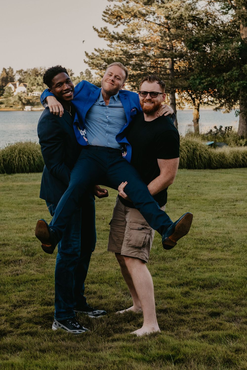 Three men laughing outdoors as two carry the middle man on a grassy lakeside lawn.