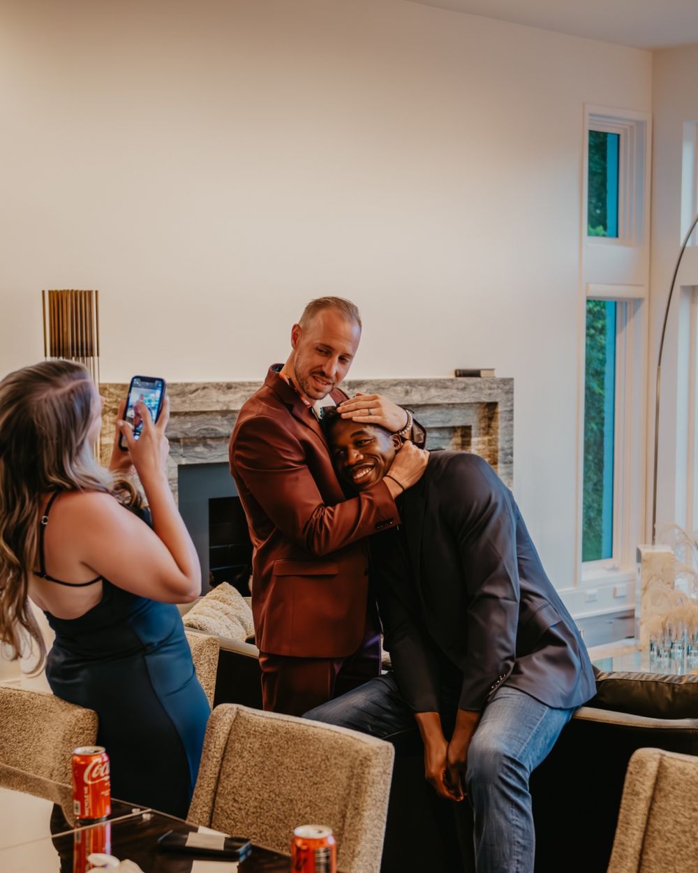 A man in a brown blazer lifts another man while a woman photographs the moment indoors.