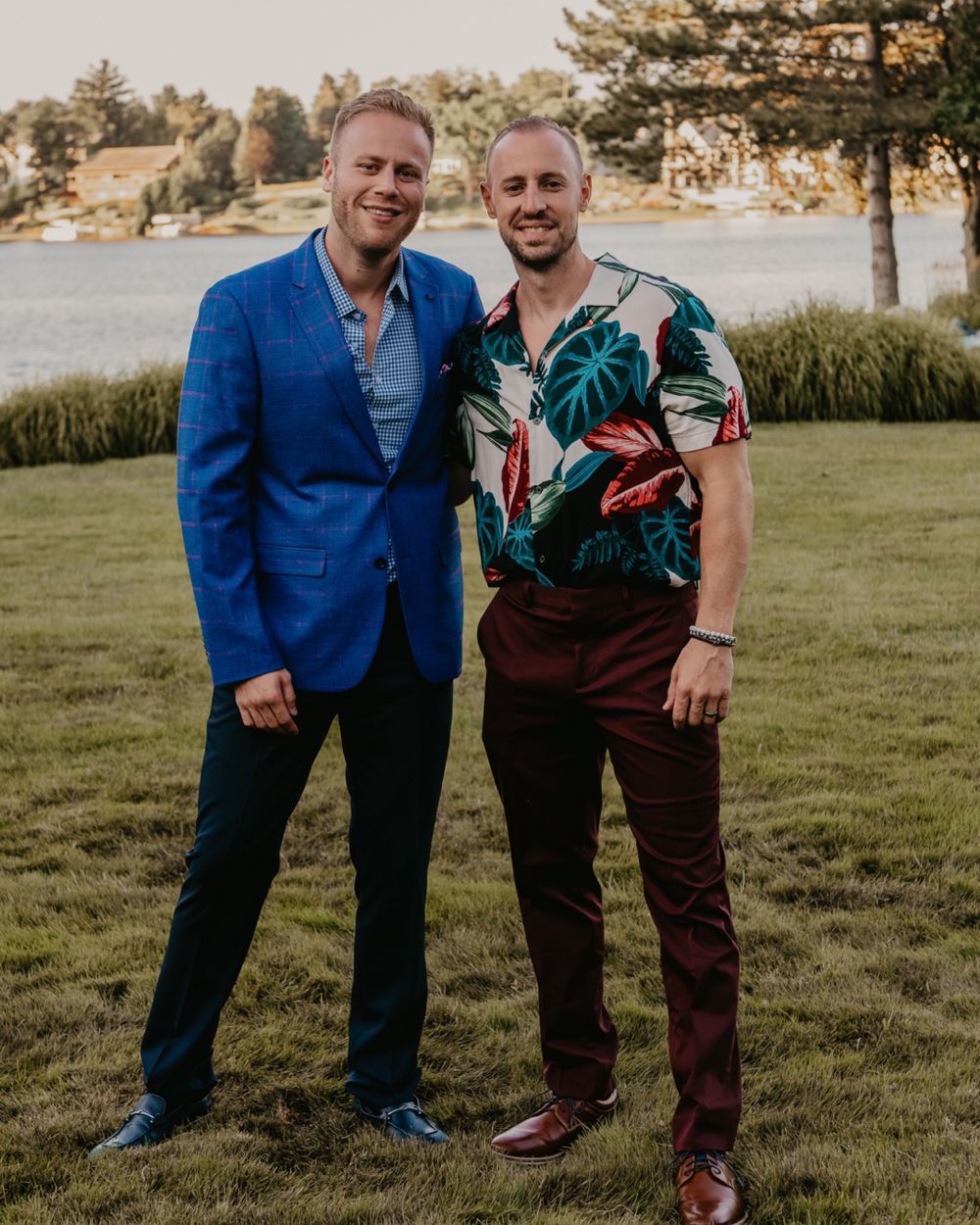 Two men smiling outdoors near a lake, one in a blue suit and one in a floral shirt.