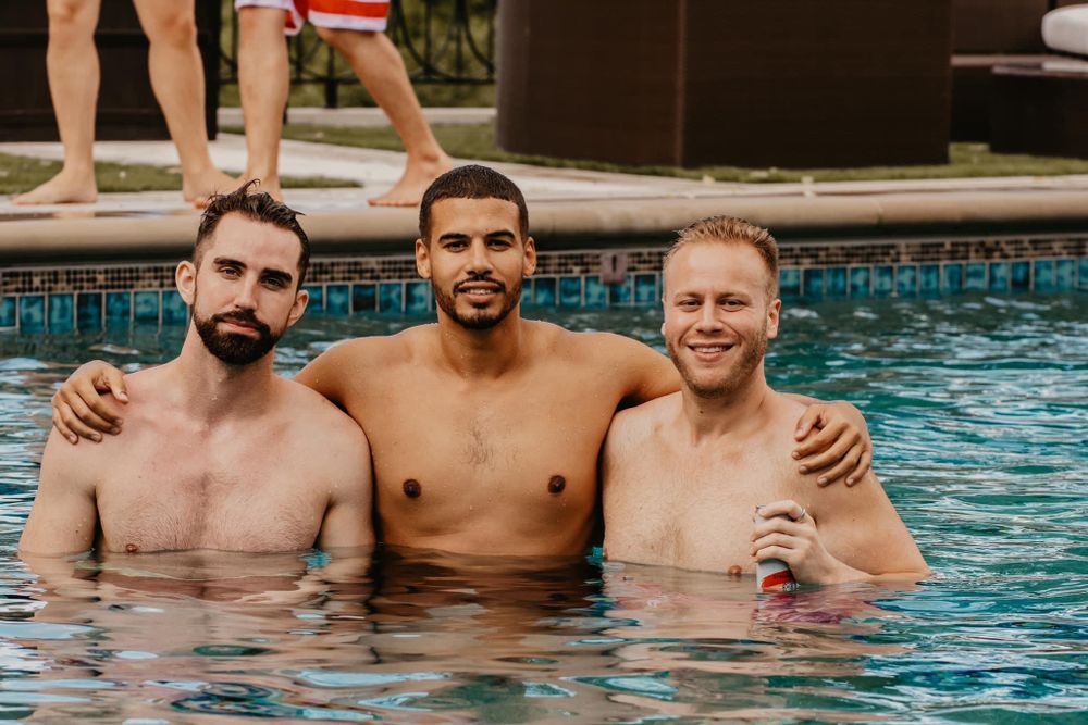 Three shirtless men smiling together in a swimming pool at an outdoor party.