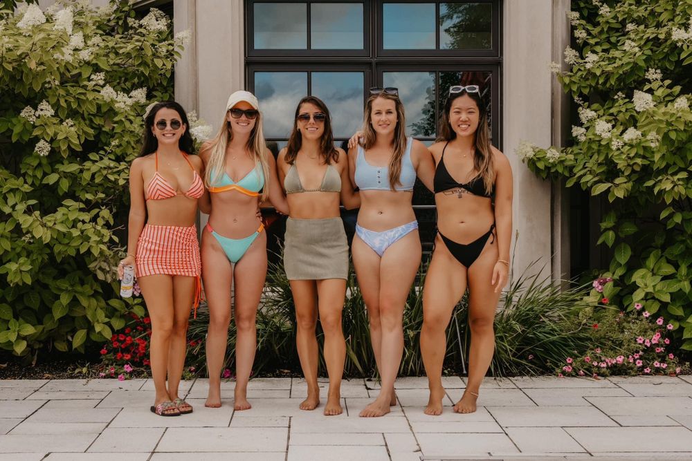 Five women in colorful swimsuits posing together in front of a floral garden wall.