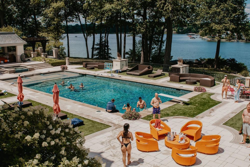 Aerial view of a luxury backyard pool party with guests near a lakeside estate.
