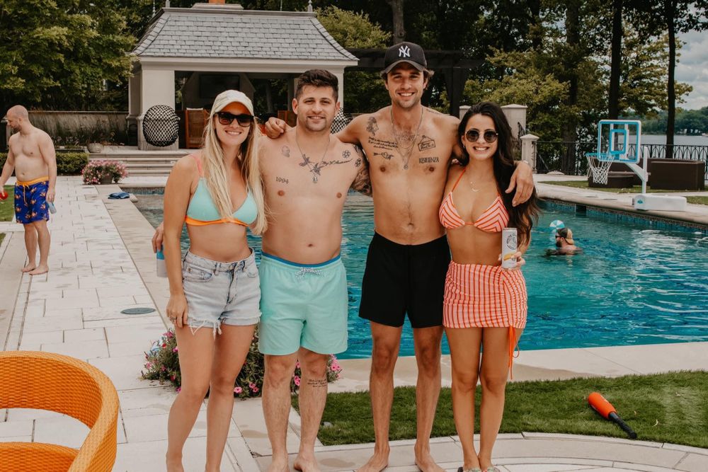 Two couples posing and smiling together beside a backyard swimming pool on a sunny day.