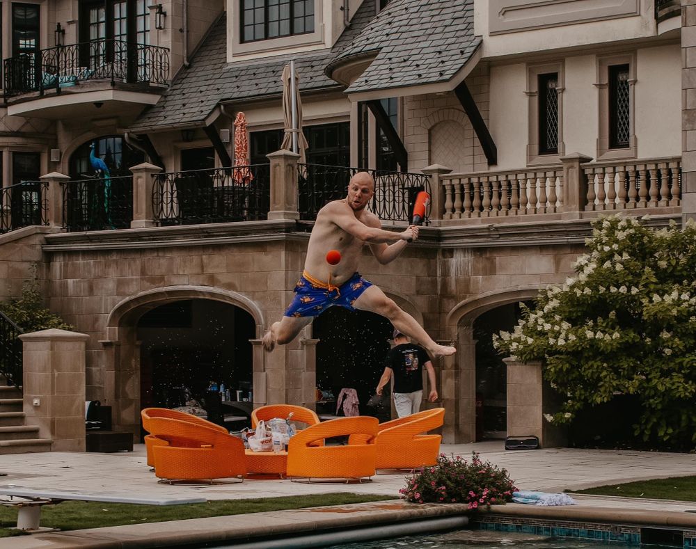 Man in blue swim trunks jumping near an orange inflatable by a luxury pool.