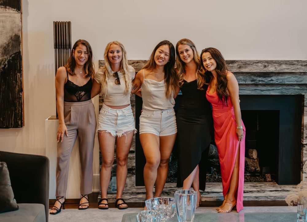 Five women smiling together in front of a stone fireplace at an indoor social event.