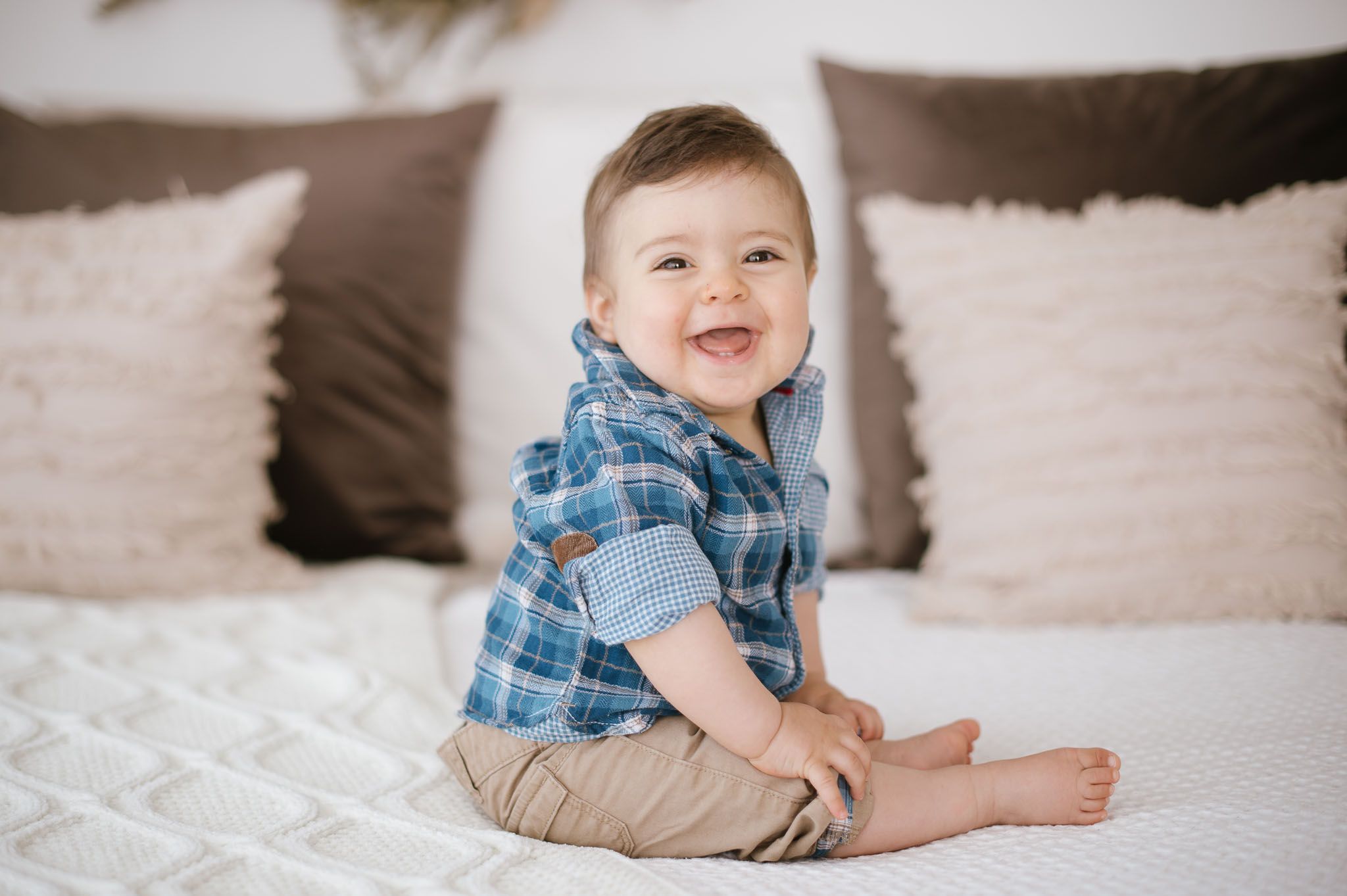 Smiling baby wearing blue plaid shirt sits on white bed with brown pillows.