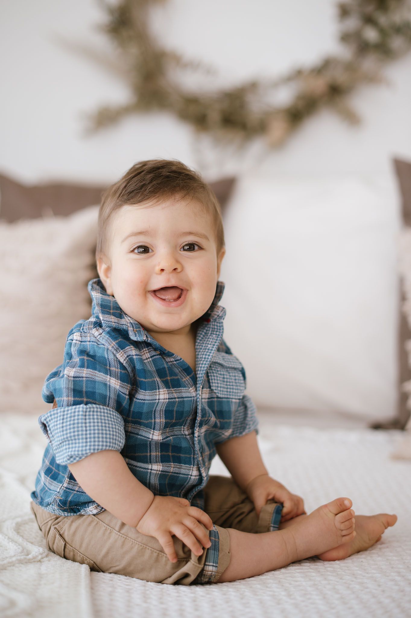 Happy baby in plaid shirt and khakis sits on white bedding with decorative wreath in background.