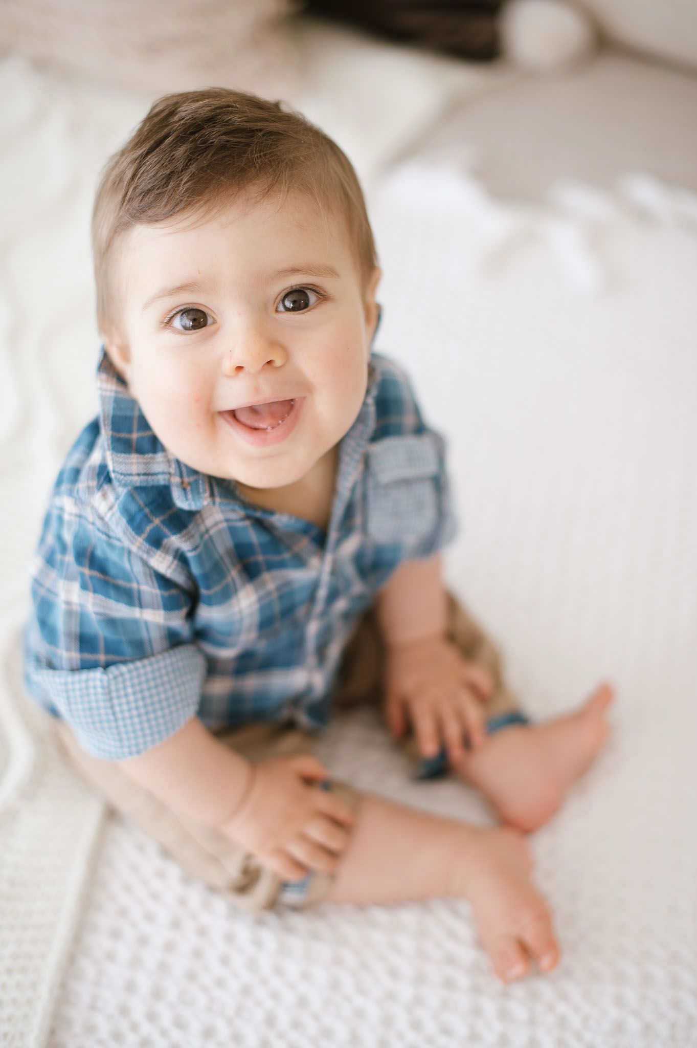 Cheerful baby in rolled-sleeve plaid shirt sits on textured white blanket.