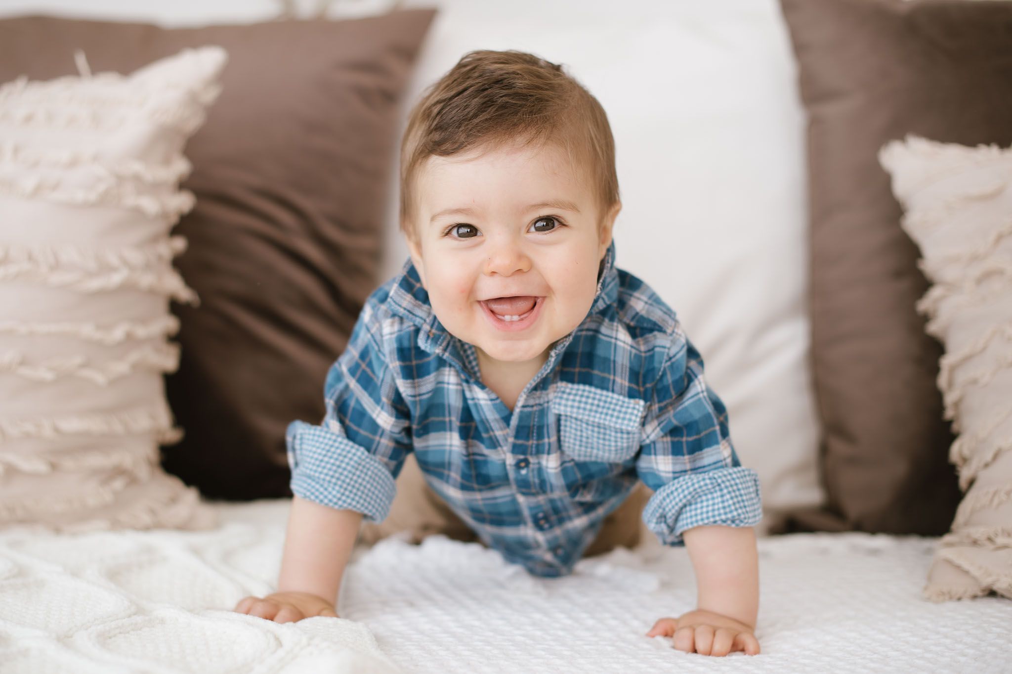 Baby in blue plaid crawls on white bedding with decorative pillows behind.
