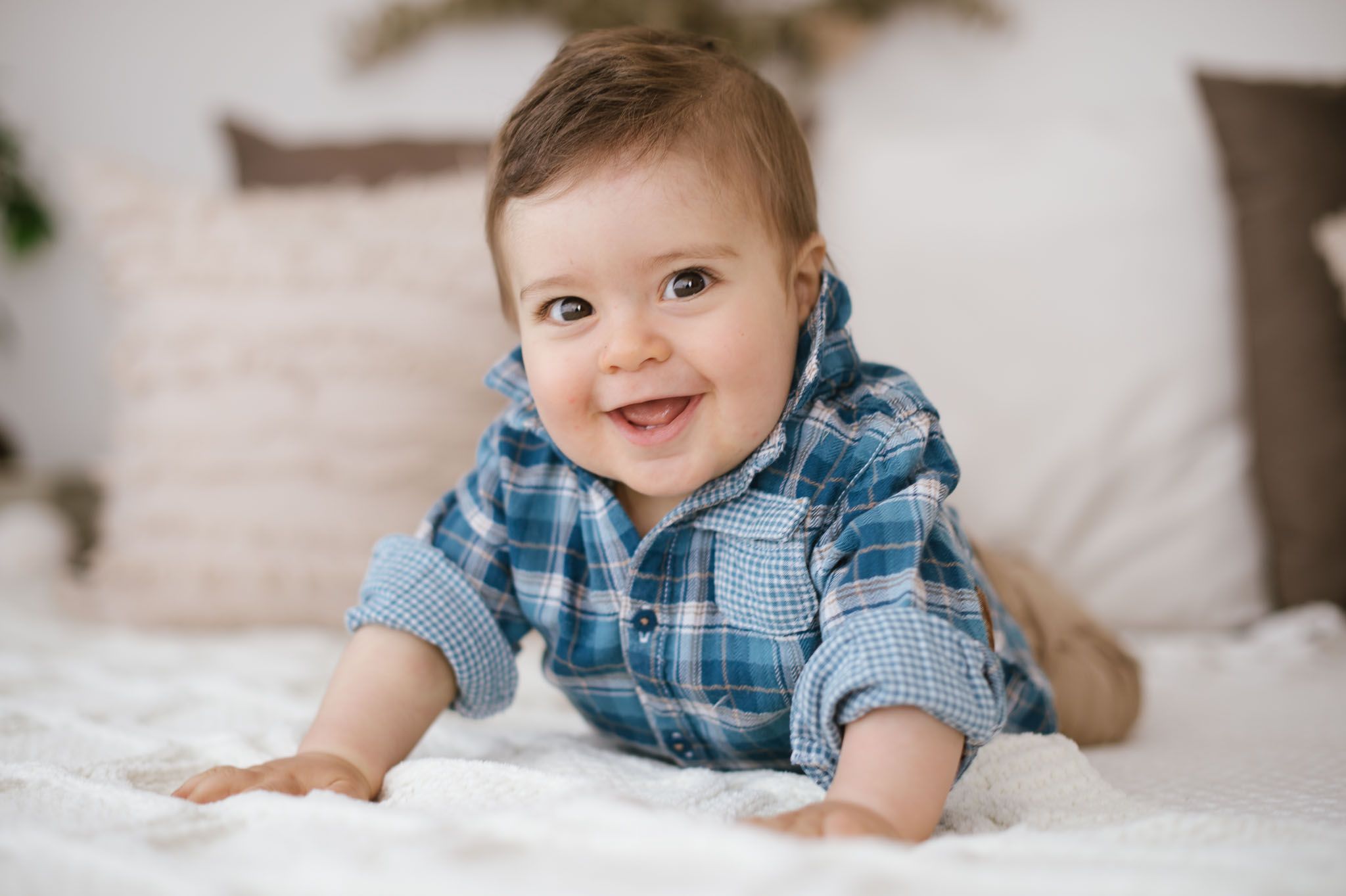 Baby in blue plaid shirt smiles brightly while lying on white bedding.