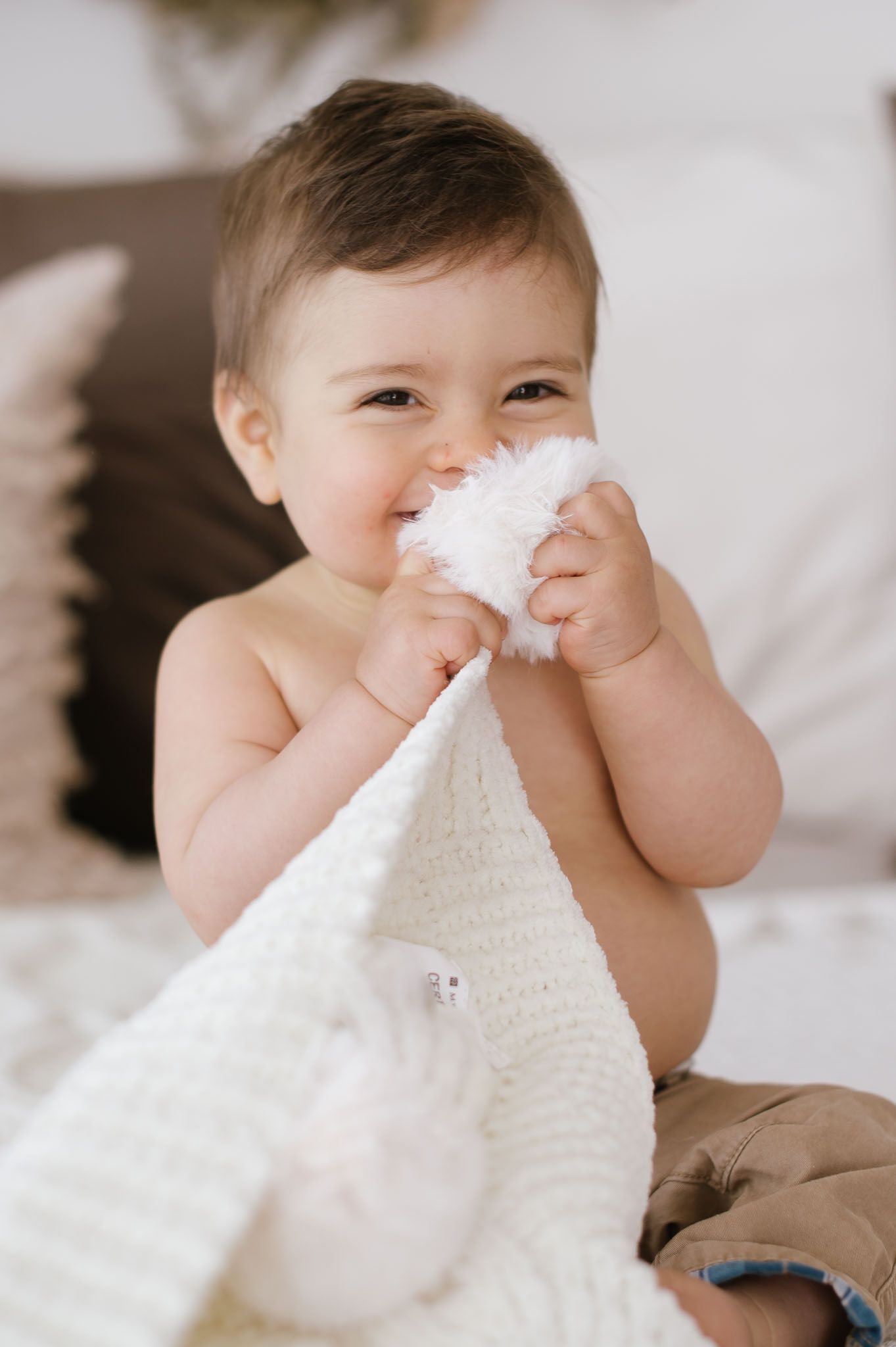 Baby sitting on bed playing with soft white blanket in cozy bedroom setting.