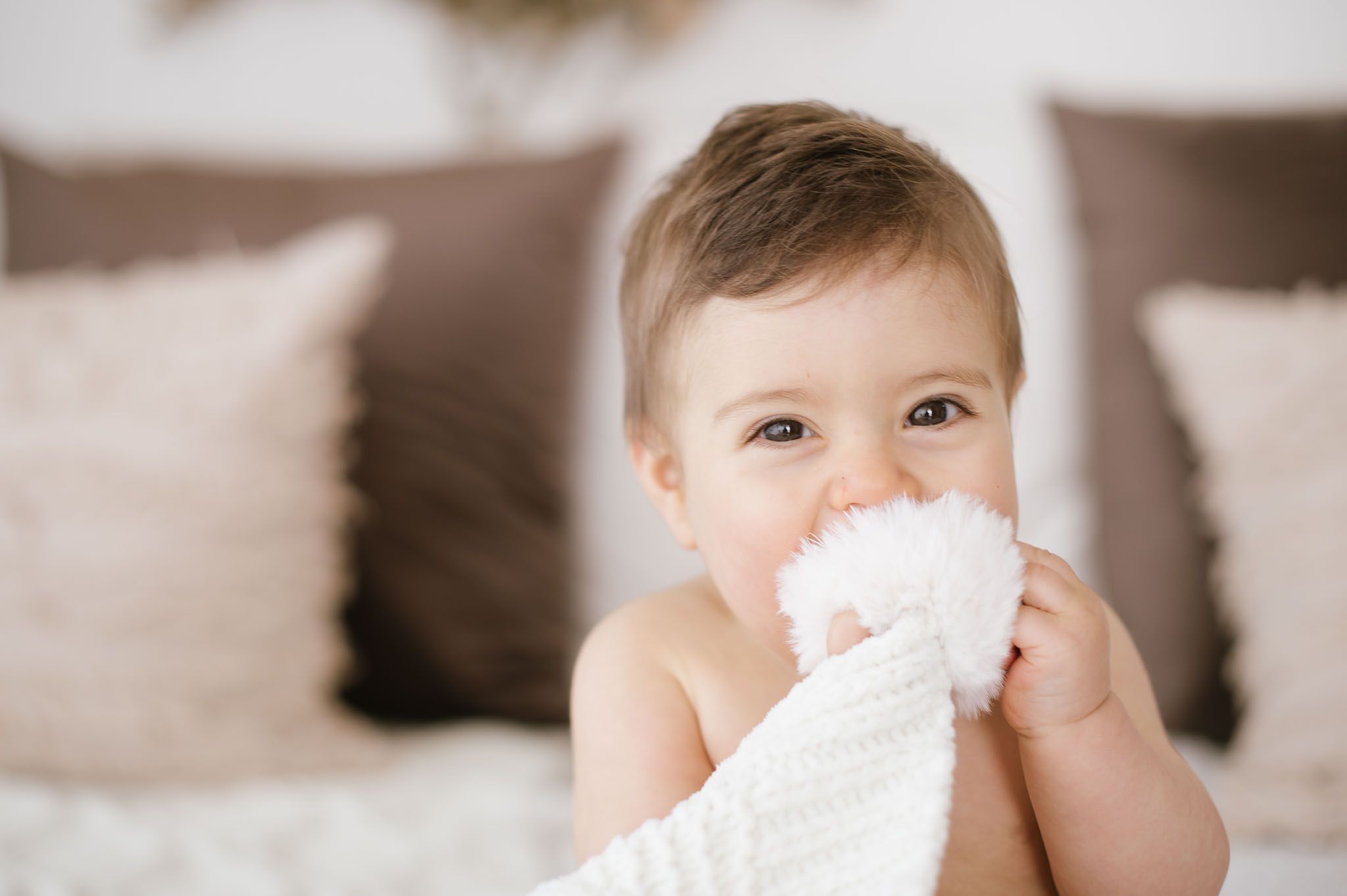 Baby sitting on bed holding white blanket near face against neutral background.