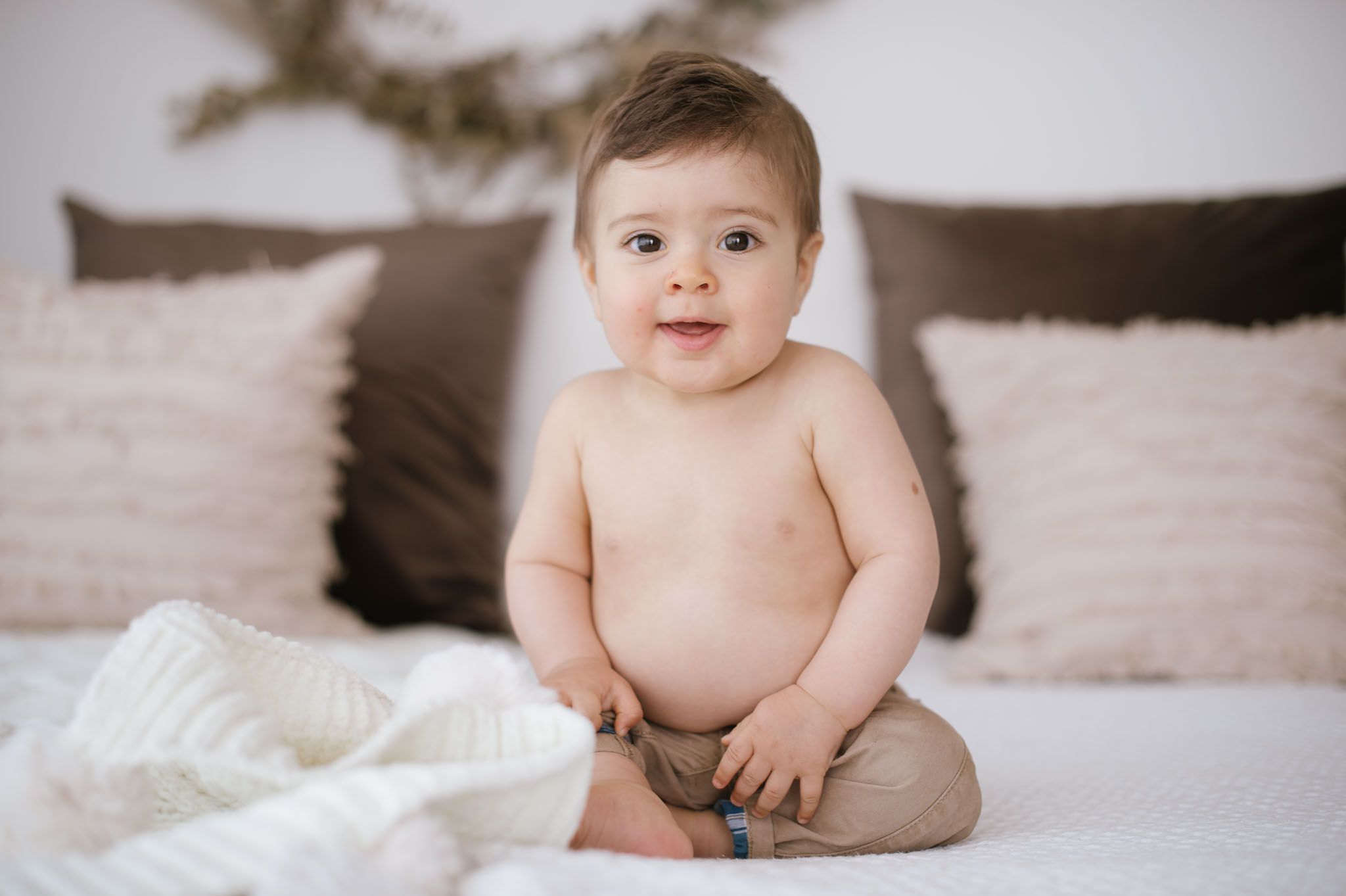 Baby sits on white bedding surrounded by beige pillows in minimalist setting.