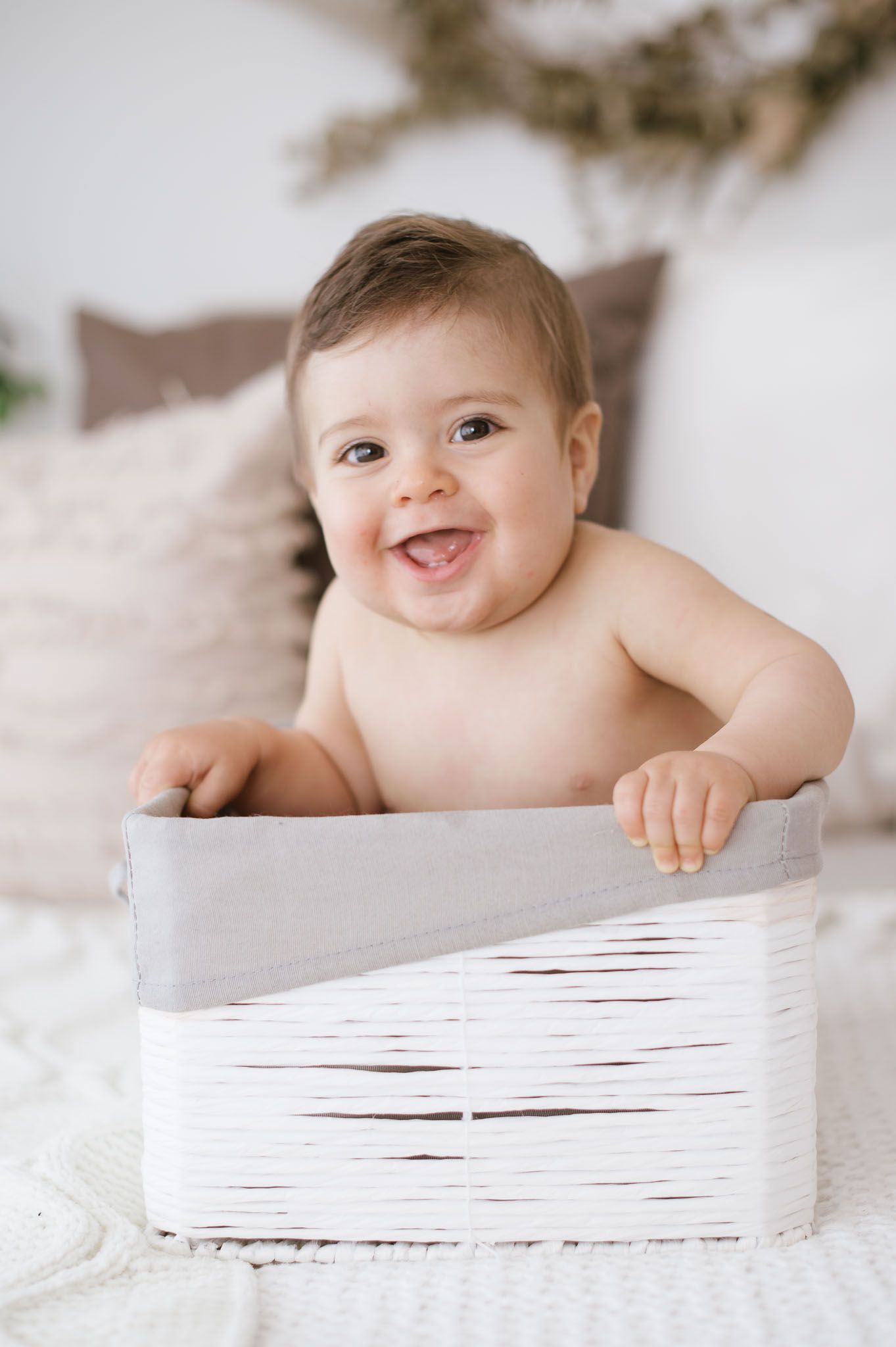 Smiling baby sitting in white decorative box on bed with neutral decor.