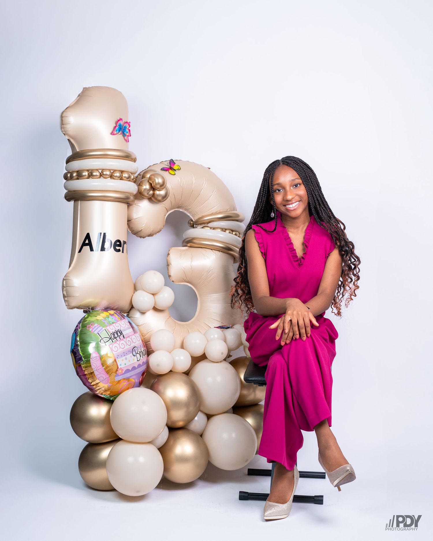 Studio portrait with gold balloon decorations and magenta jumpsuit against white background.