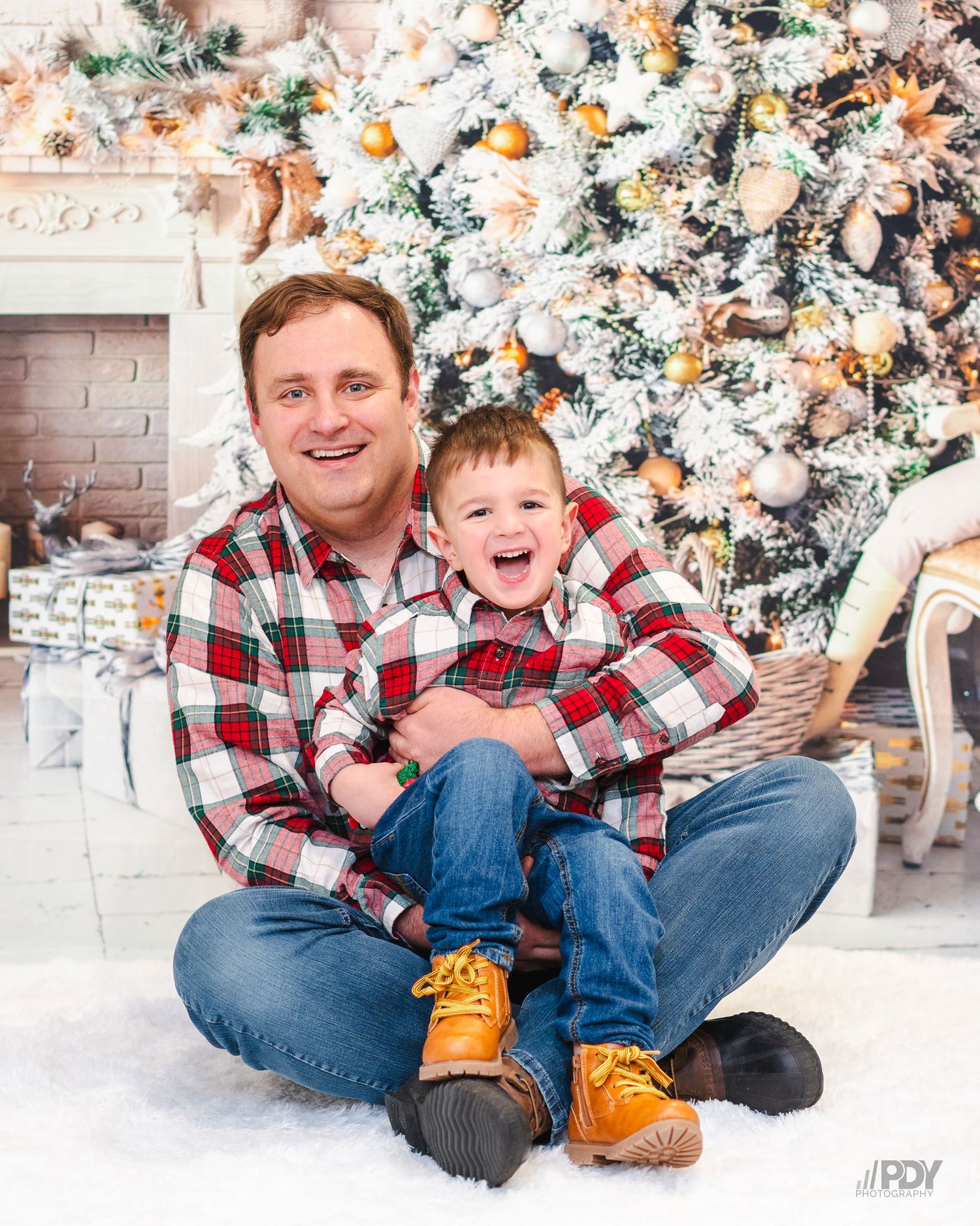Child in red plaid shirt sits on floor near festive Christmas decorations.