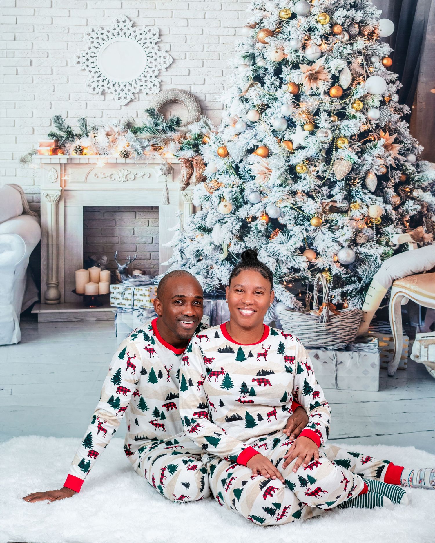 Couple in matching holiday pajamas sitting by decorated Christmas tree and fireplace.