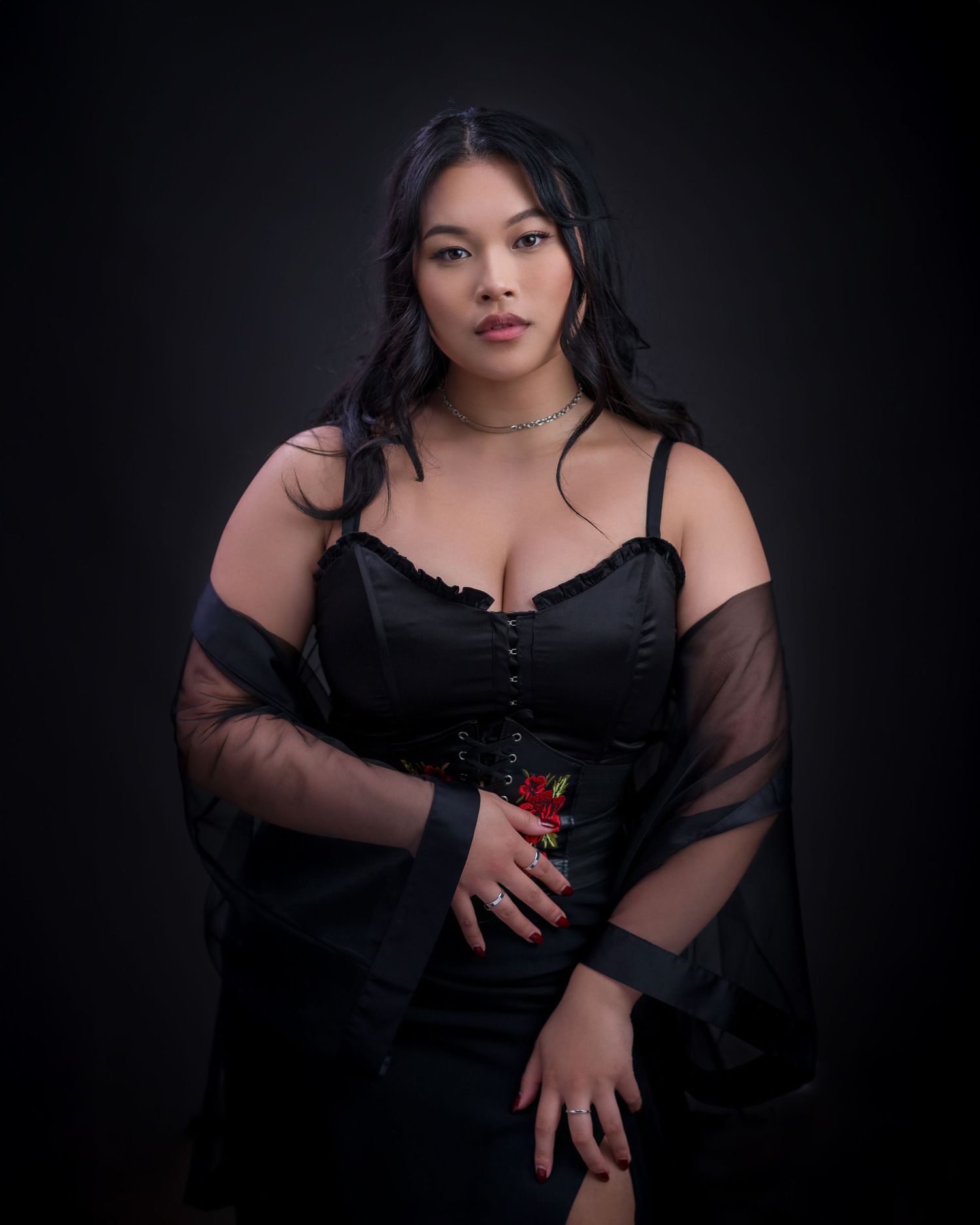 Dramatic studio portrait in black sheer-sleeved dress against dark background.