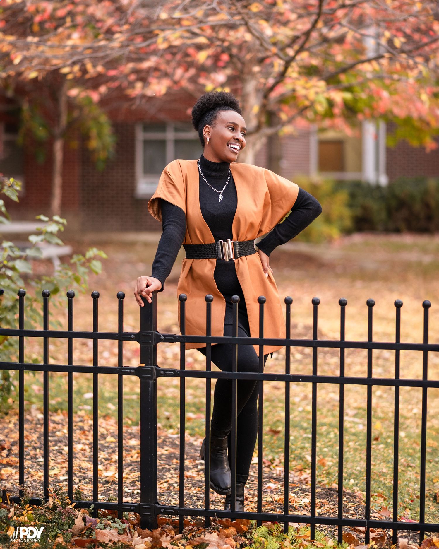 Fashion model in orange vest and black outfit poses confidently by iron fence during autumn season.
