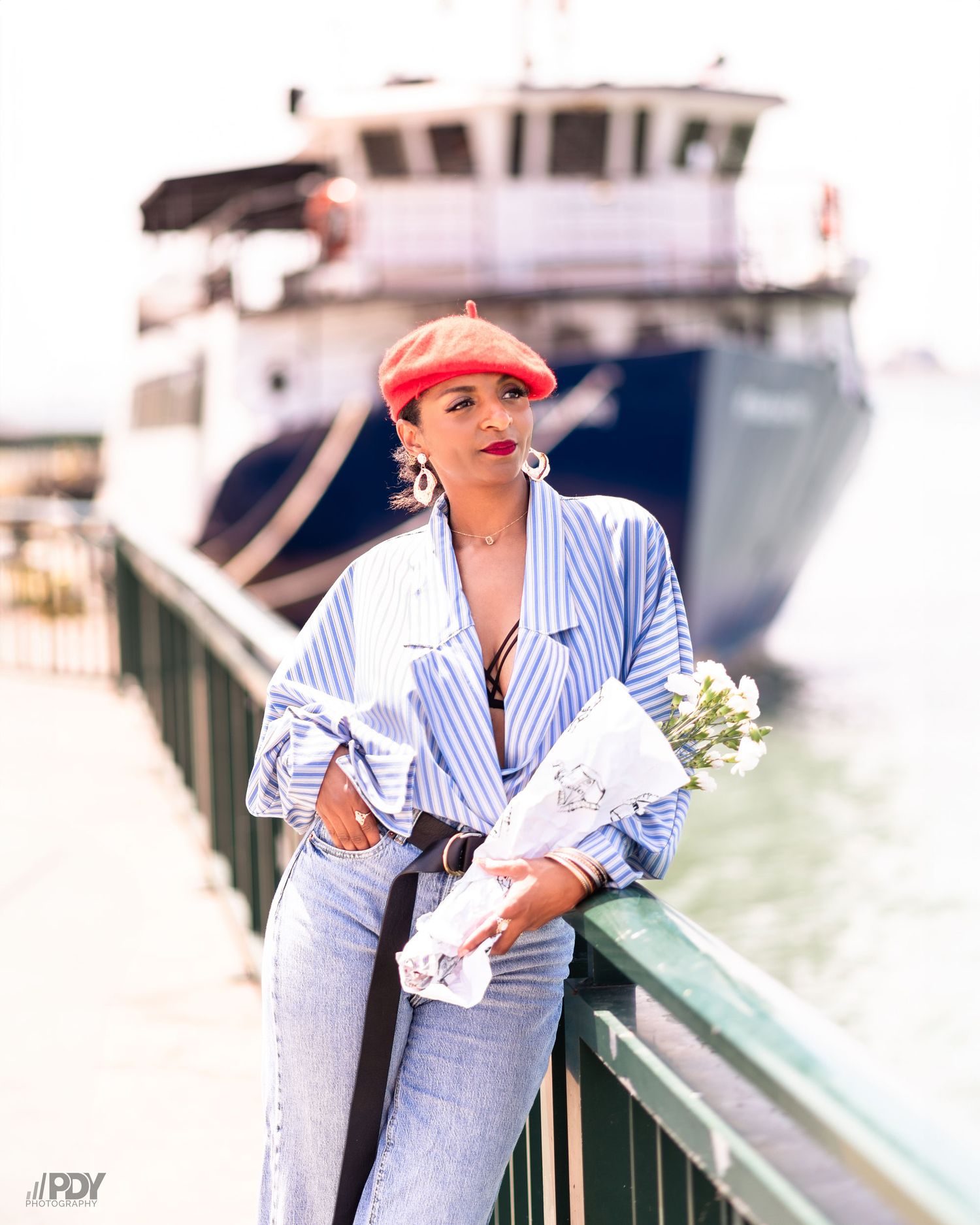 Casual maritime style with striped button-down and red cap near large vessel.