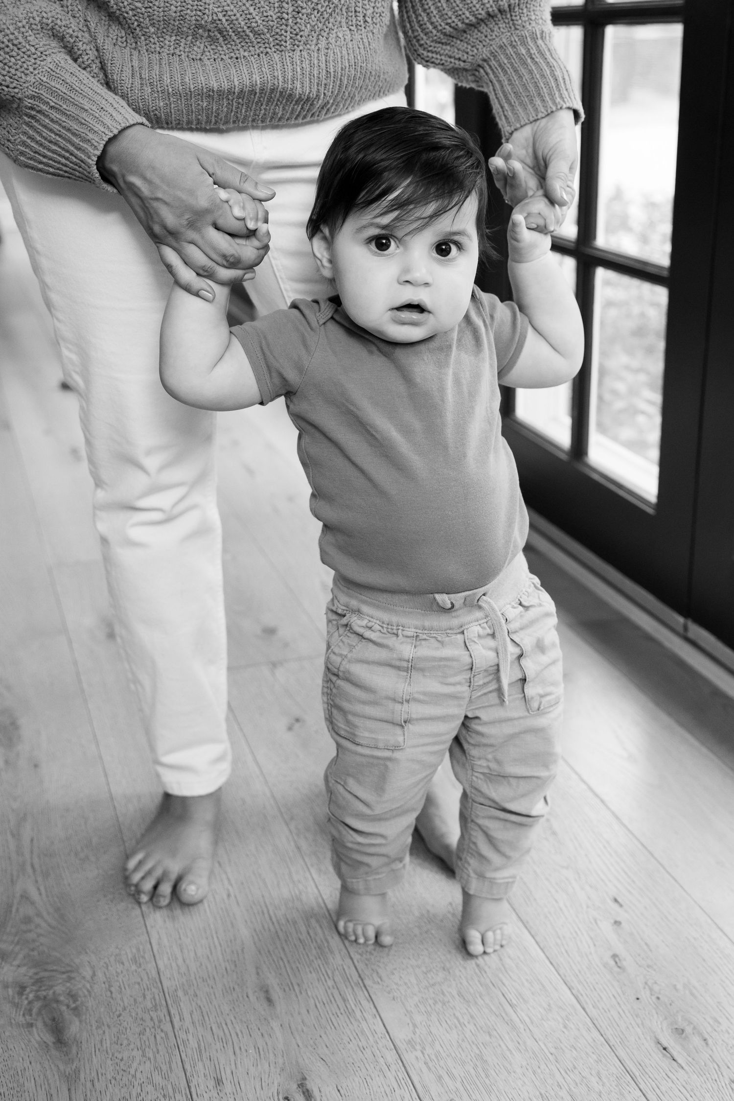 Black and white photo of a curious baby taking first steps while holding an adult's hands indoors.