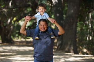 Happy grandfather carrying young grandson on his shoulders while walking through a tree-lined path.
