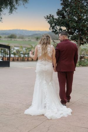 Bride in lace wedding gown and groom in burgundy suit walking hand in hand at sunset.