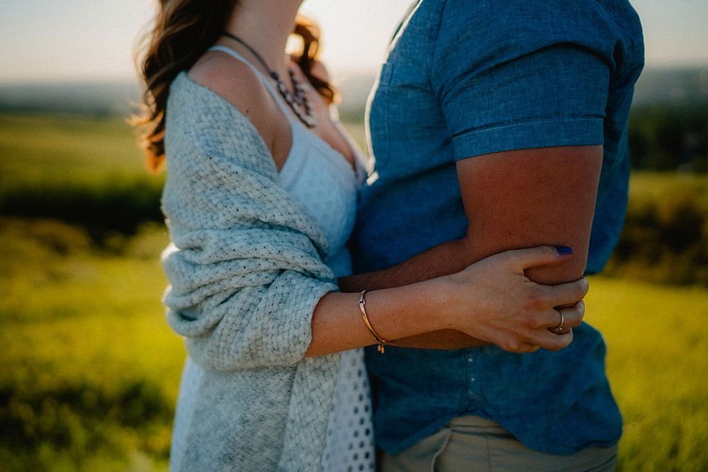 Close up of couple in casual attire embracing in golden sunlight on grassy hillside.
