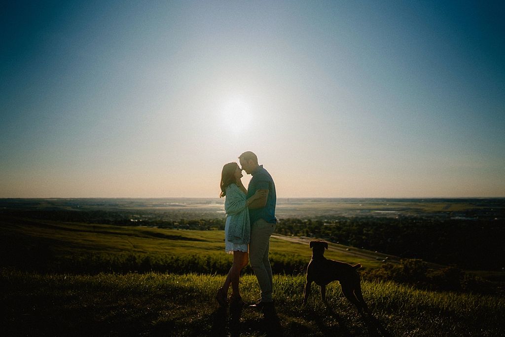 Silhouetted couple stand with their dog against setting sun on hilltop landscape.