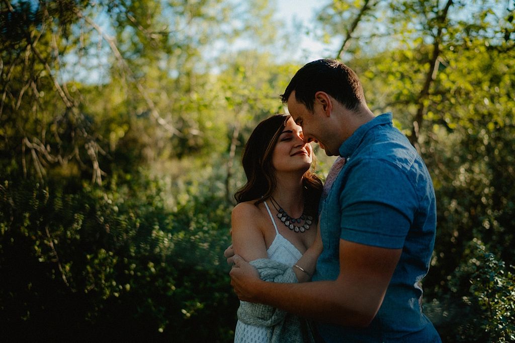 A romantic couple embraces on a sunlit forest path surrounded by lush green trees.