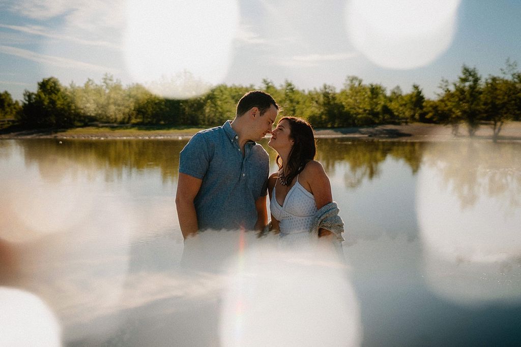 Couple embracing by a sunlit lake with trees in the background during a summer afternoon.