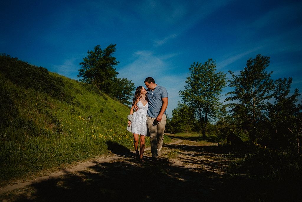A couple walks together along a countryside path surrounded by green hills and trees under a deep blue sky.