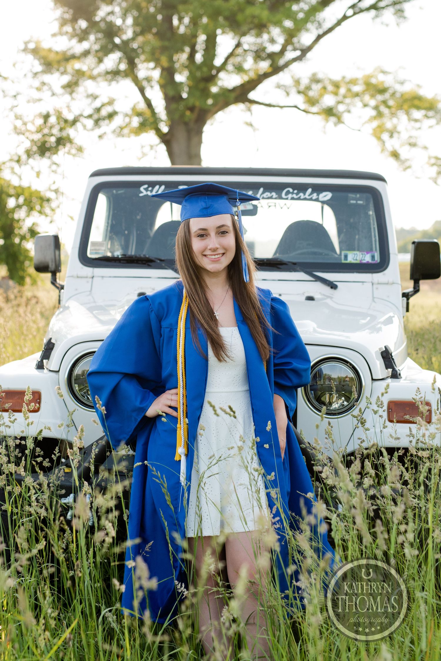 Graduate in blue cap and gown poses in wildflower field with white Jeep in background.